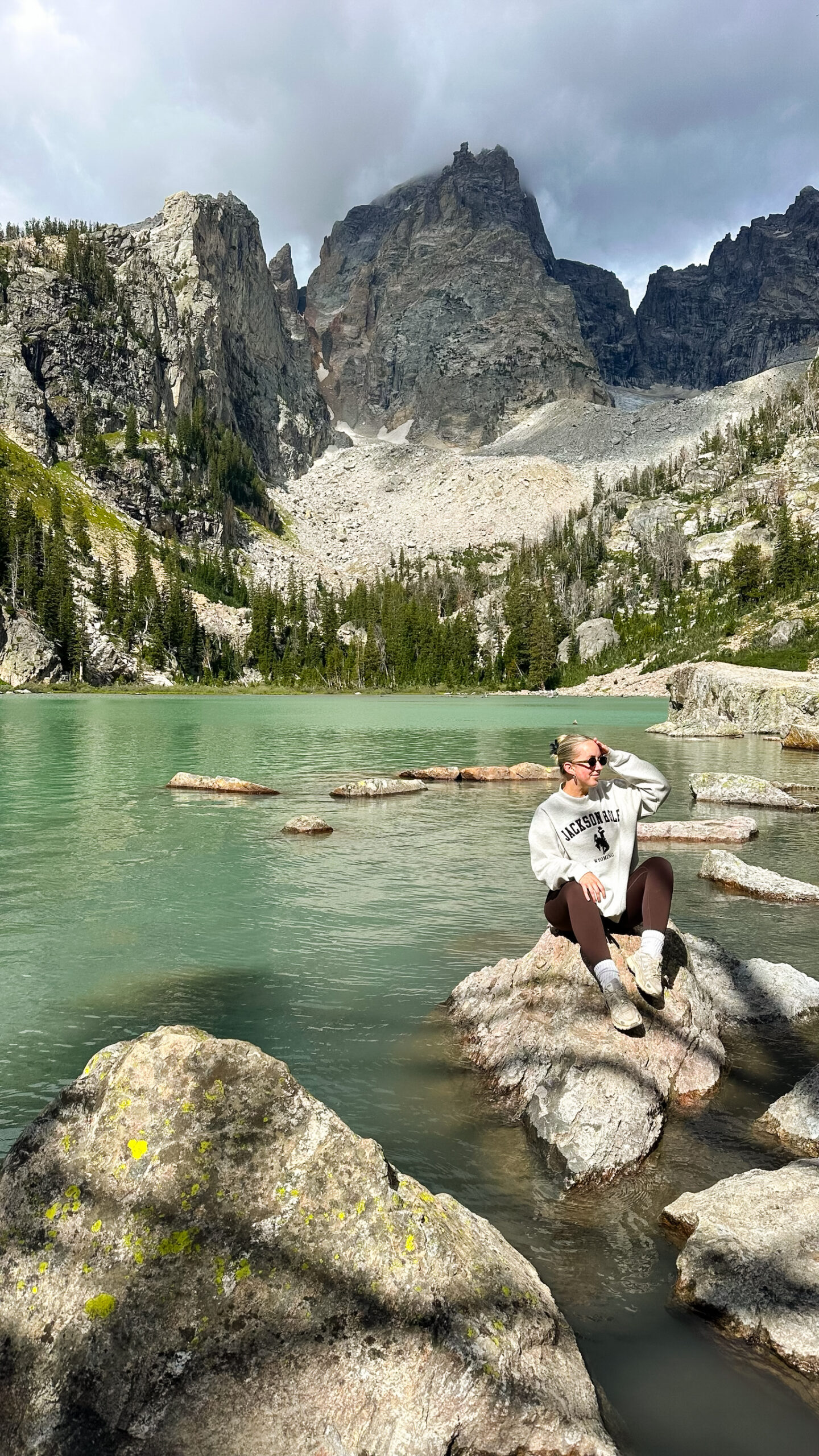 Woman in a Jackson Hole sweatshirt sitting on a rock at the turquoise waters of Delta Lake in Grand Teton National Park