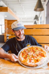 Guest smiling over a fresh pepperoni jalapeño pizza at Oven and Tap in downtown Bentonville, Arkansas, a favorite rehearsal dinner spot