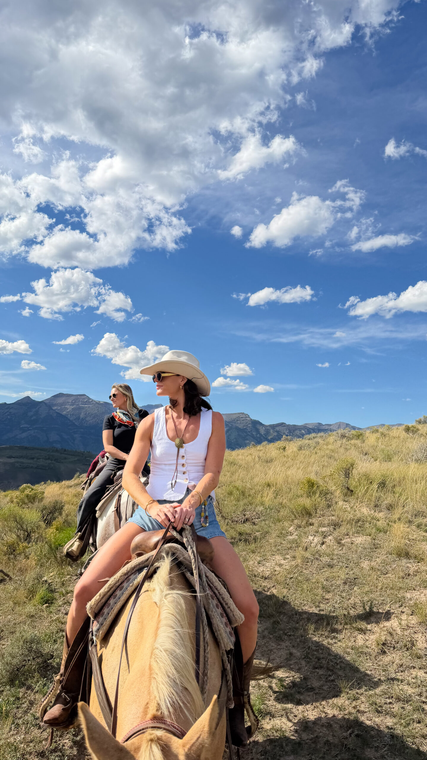 Two women on horseback riding through sagebrush hills with the Grand Teton mountains rising behind them near Jackson Hole, Wyoming